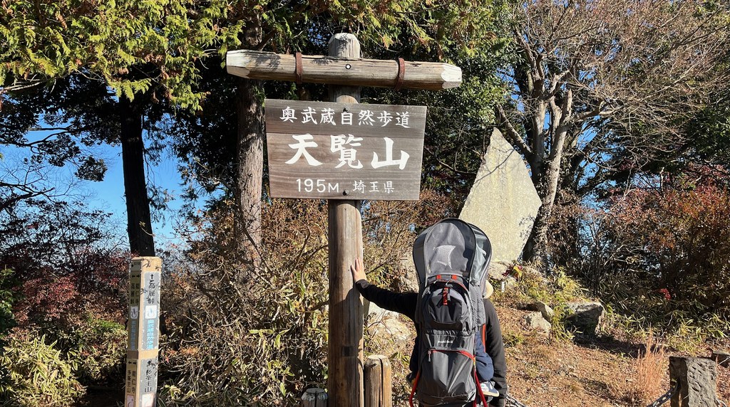 天覧山の山頂の標識と、子どもを背負う登山者